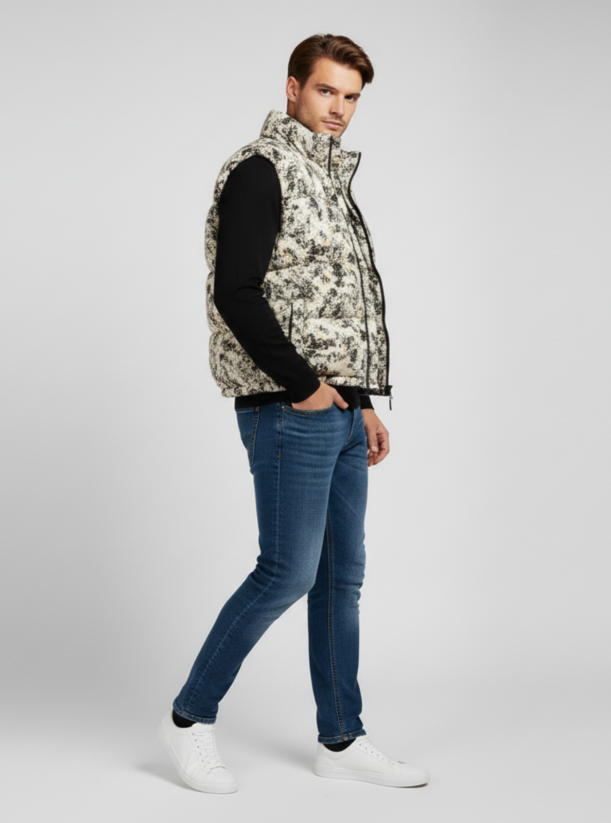 Man wearing a patterned vest, black long-sleeve shirt, blue jeans, and white sneakers on a light gray background