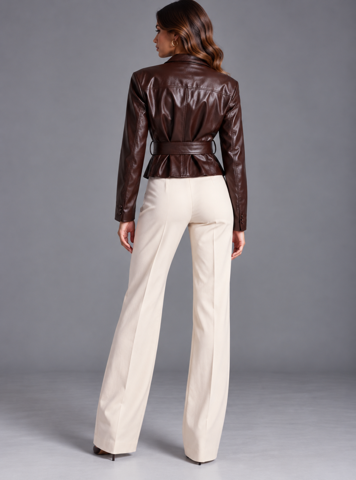 A woman shown from the back wearing a dark brown faux-leather belted jacket and cream-colored wide-leg trousers, standing against a neutral gray studio background.