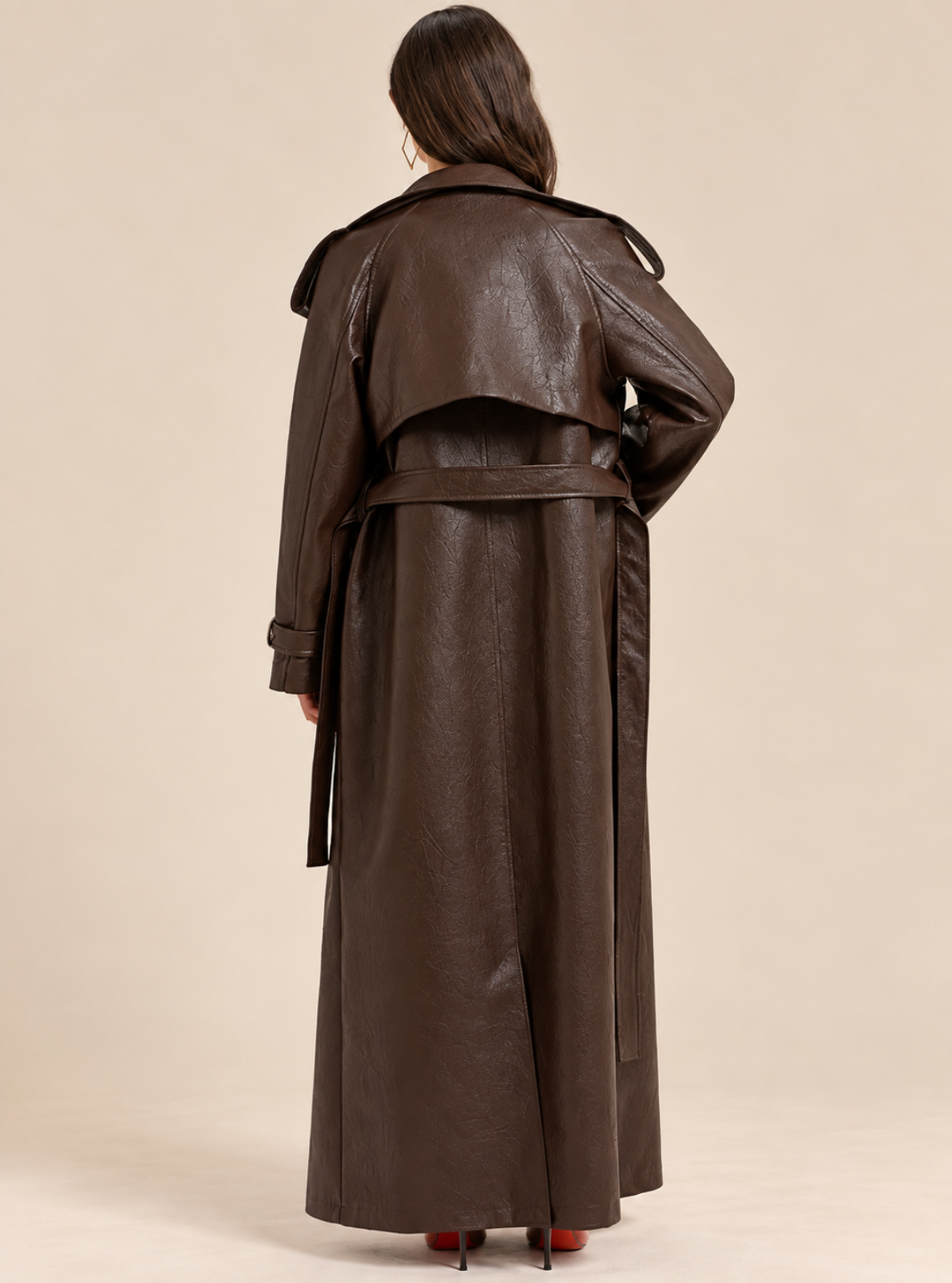 A model shown from the back wearing a long, dark brown faux-leather trench coat with a matching belt, set against a solid beige studio background.