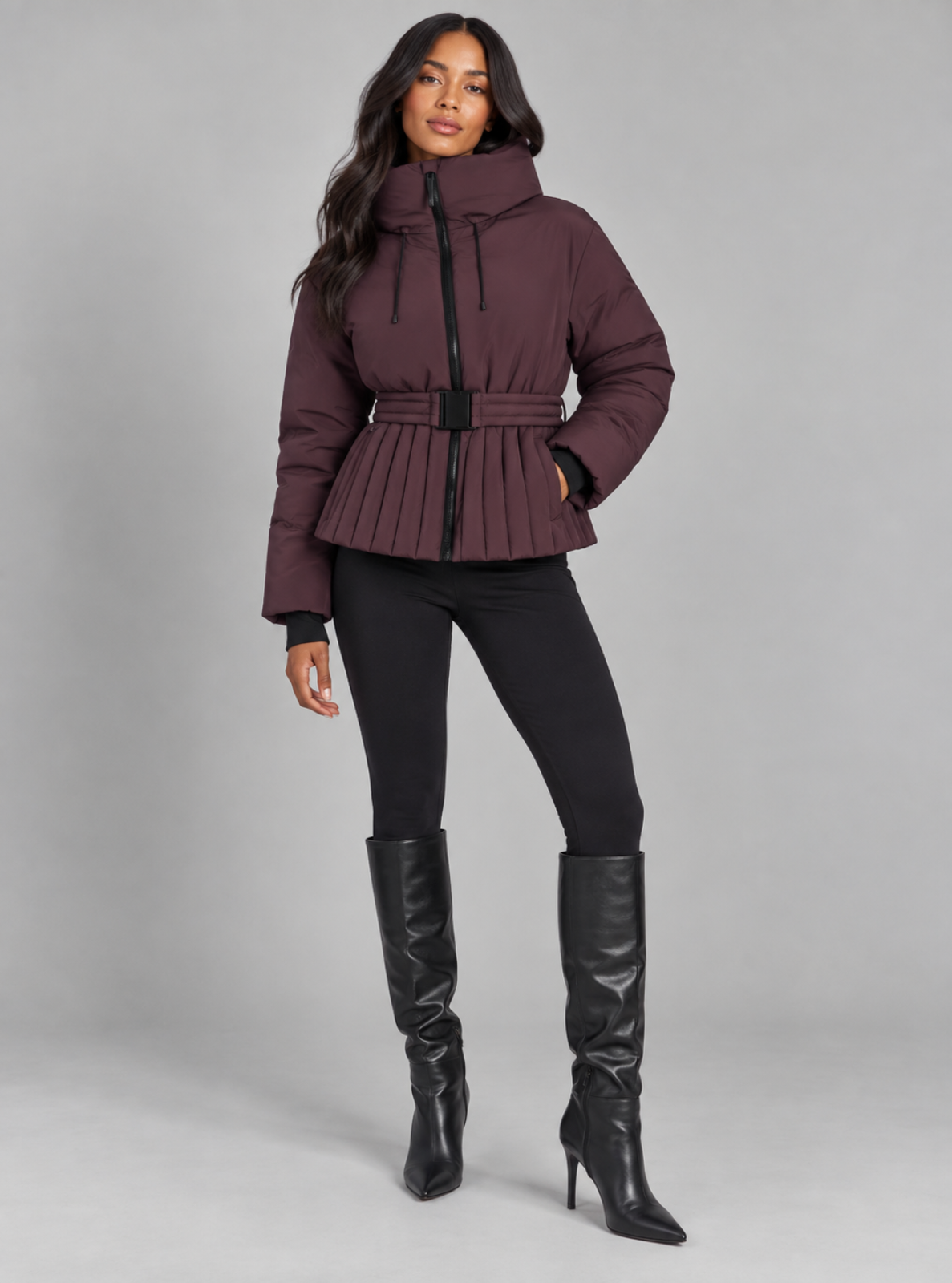 A woman models a dark plum-brown belted puffer jacket with black trim, paired with black pants and tall black boots, against a solid gray studio background.