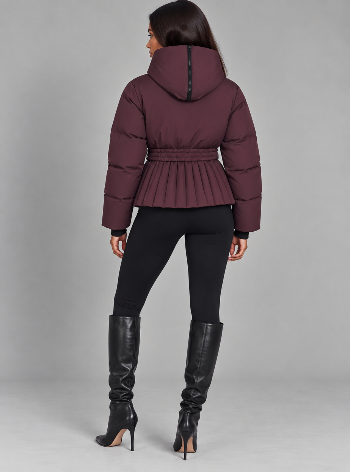 A woman shown from the back wearing a dark plum-burgundy hooded puffer jacket with a pleated waist, black leggings, and black knee-high heeled boots against a light gray studio background.