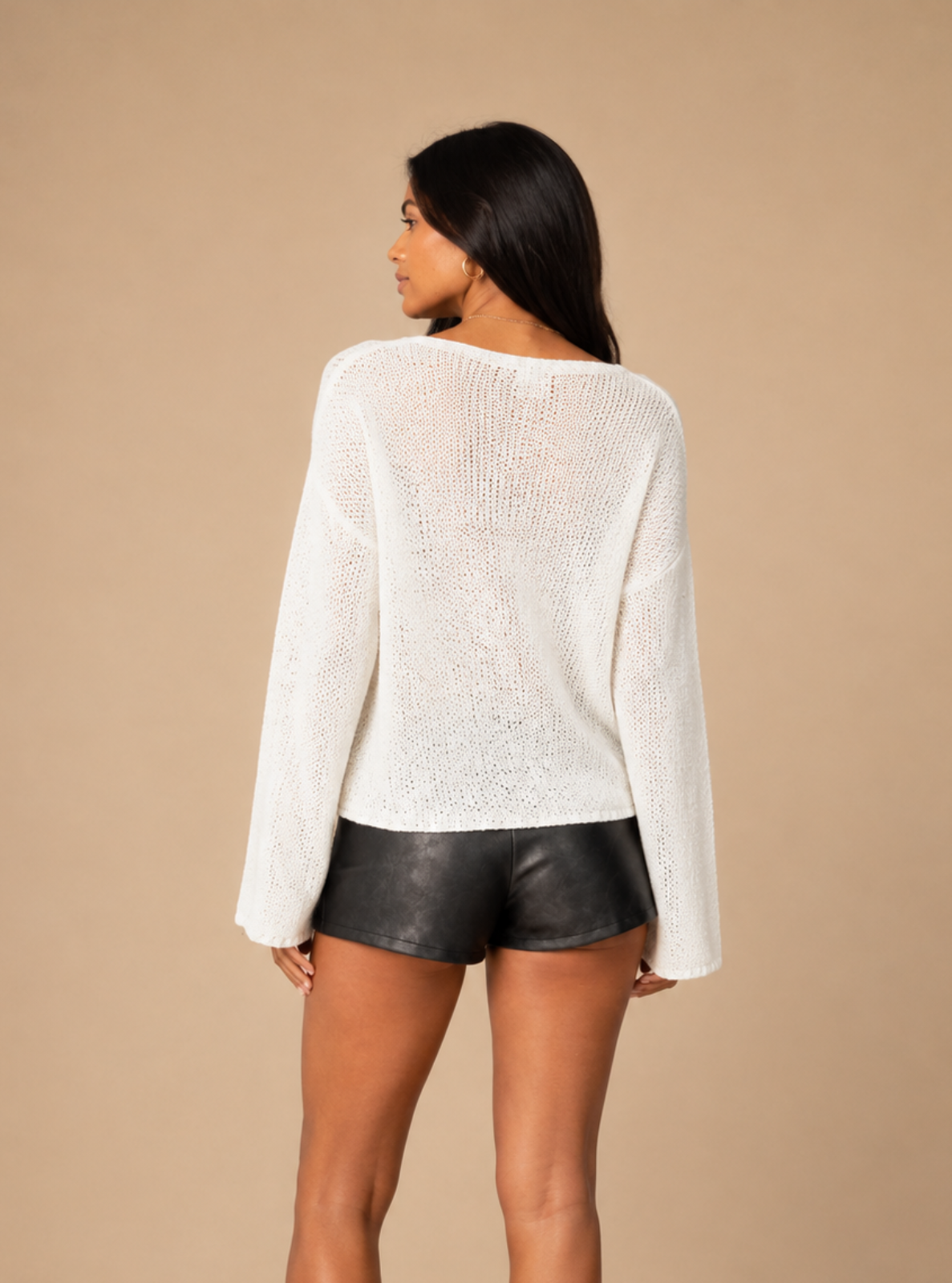 A woman is shown from the back wearing a white long-sleeve knit top and black faux-leather shorts against a solid beige studio background.