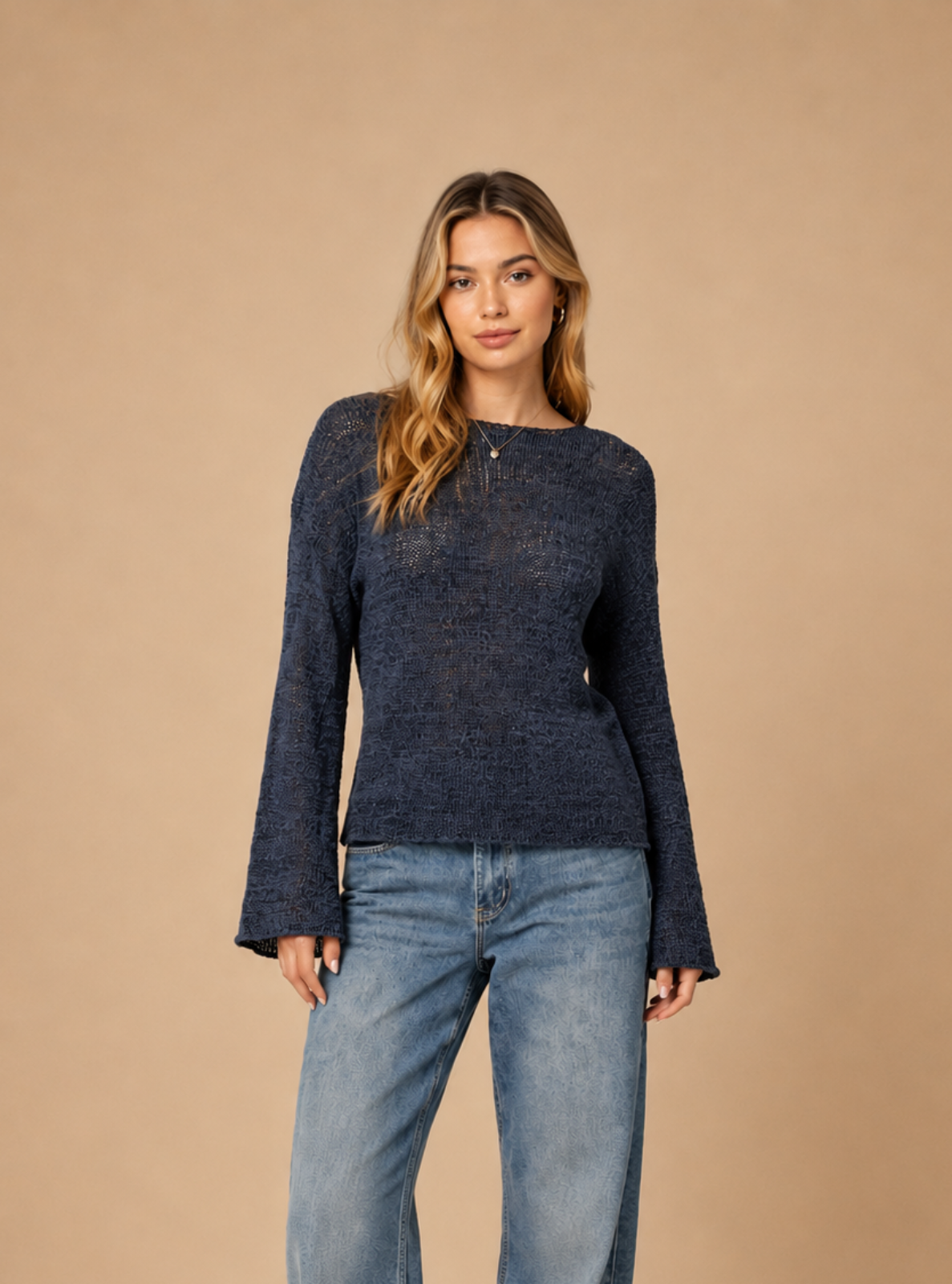 A woman modeling a dark navy blue long-sleeve knit top with wide-leg blue jeans and black sandals, standing against a solid beige studio background.