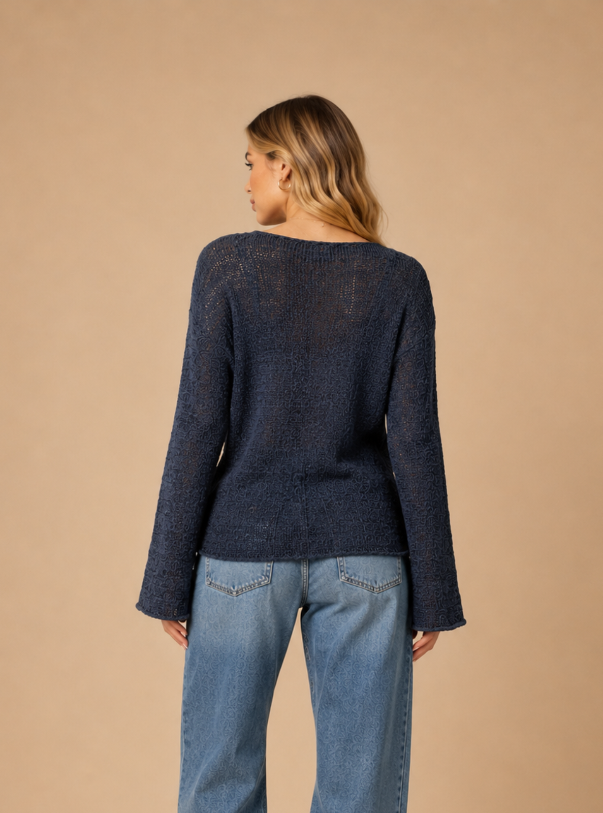 A woman is shown from the back wearing a dark navy blue long-sleeve knit sweater and wide-leg light blue jeans, standing against a solid beige studio background.