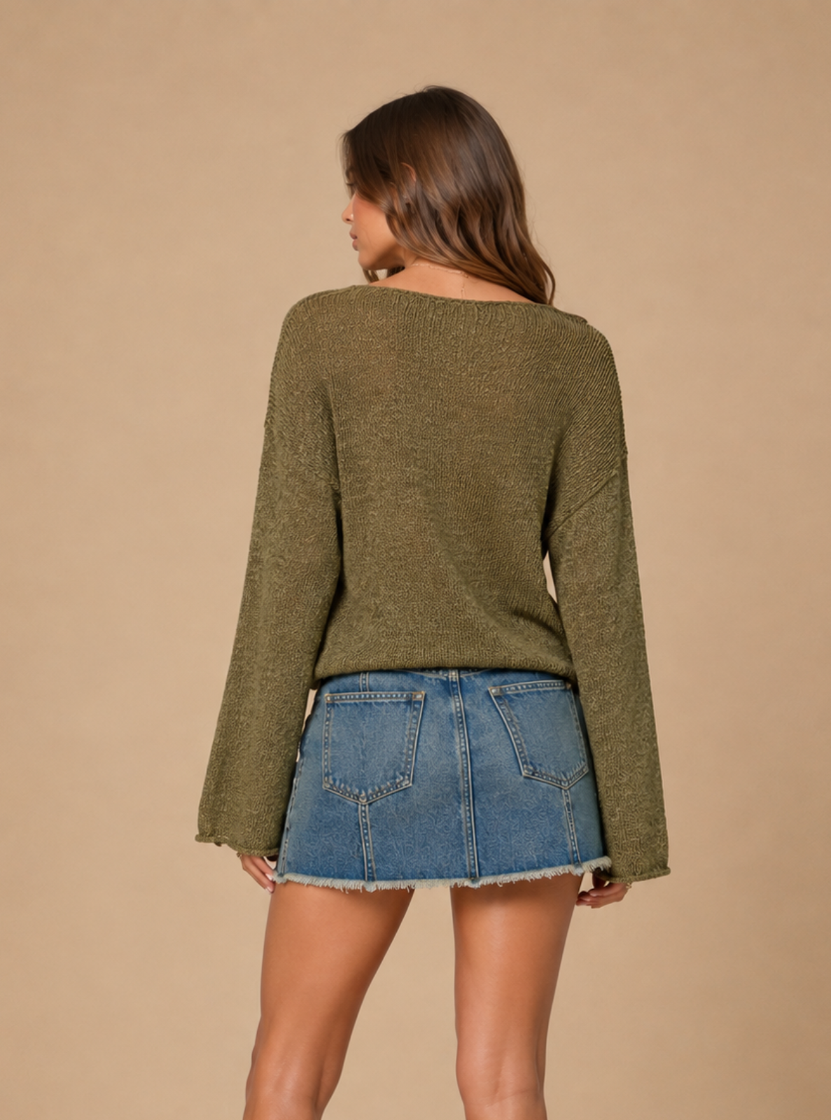 A woman is shown from the back wearing an olive green long-sleeve knit sweater and a blue denim mini skirt, standing against a solid beige studio background.