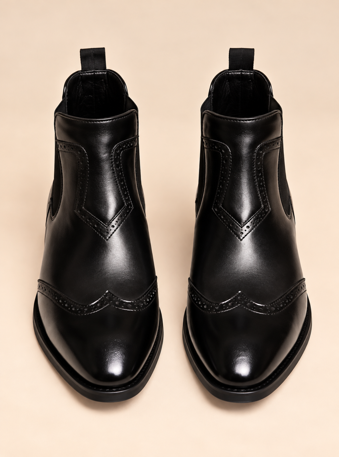 A pair of black leather Chelsea-style ankle boots with decorative brogue detailing on the toe, shown from a top-down view against a solid beige background.