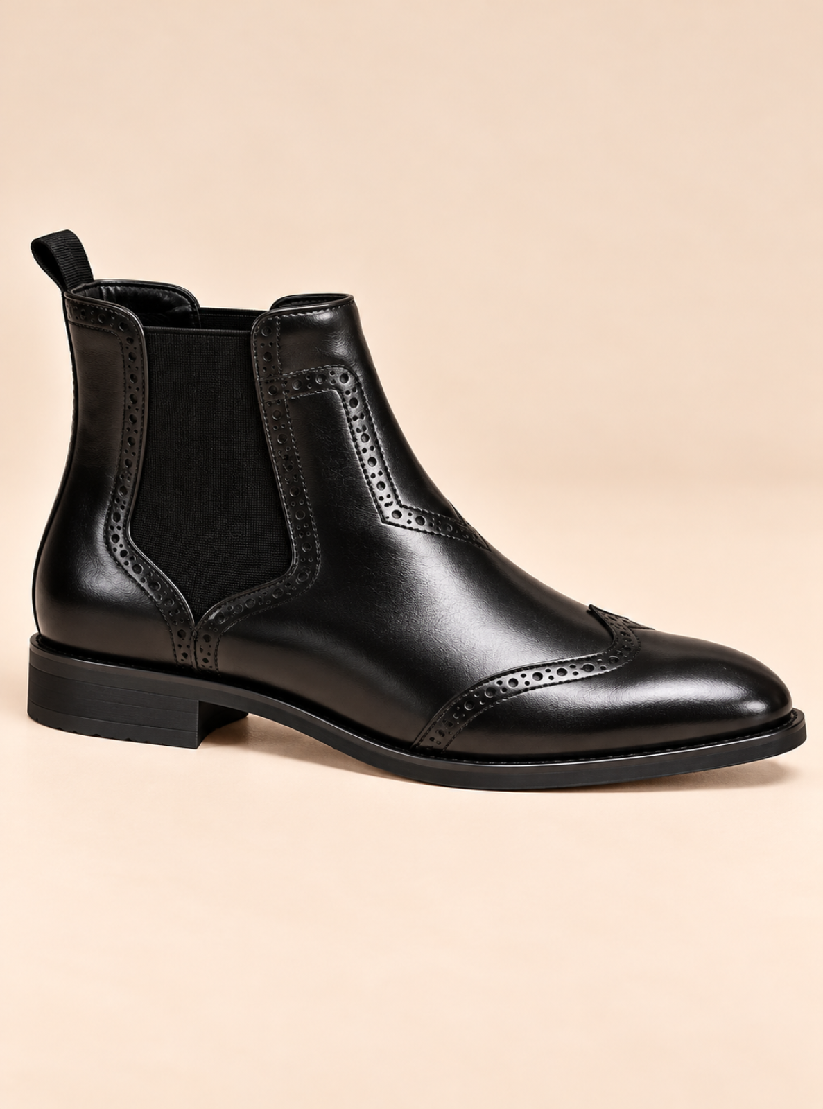 A black leather Chelsea boot with brogue detailing and a black elastic side panel, shown from a side profile against a solid beige background.