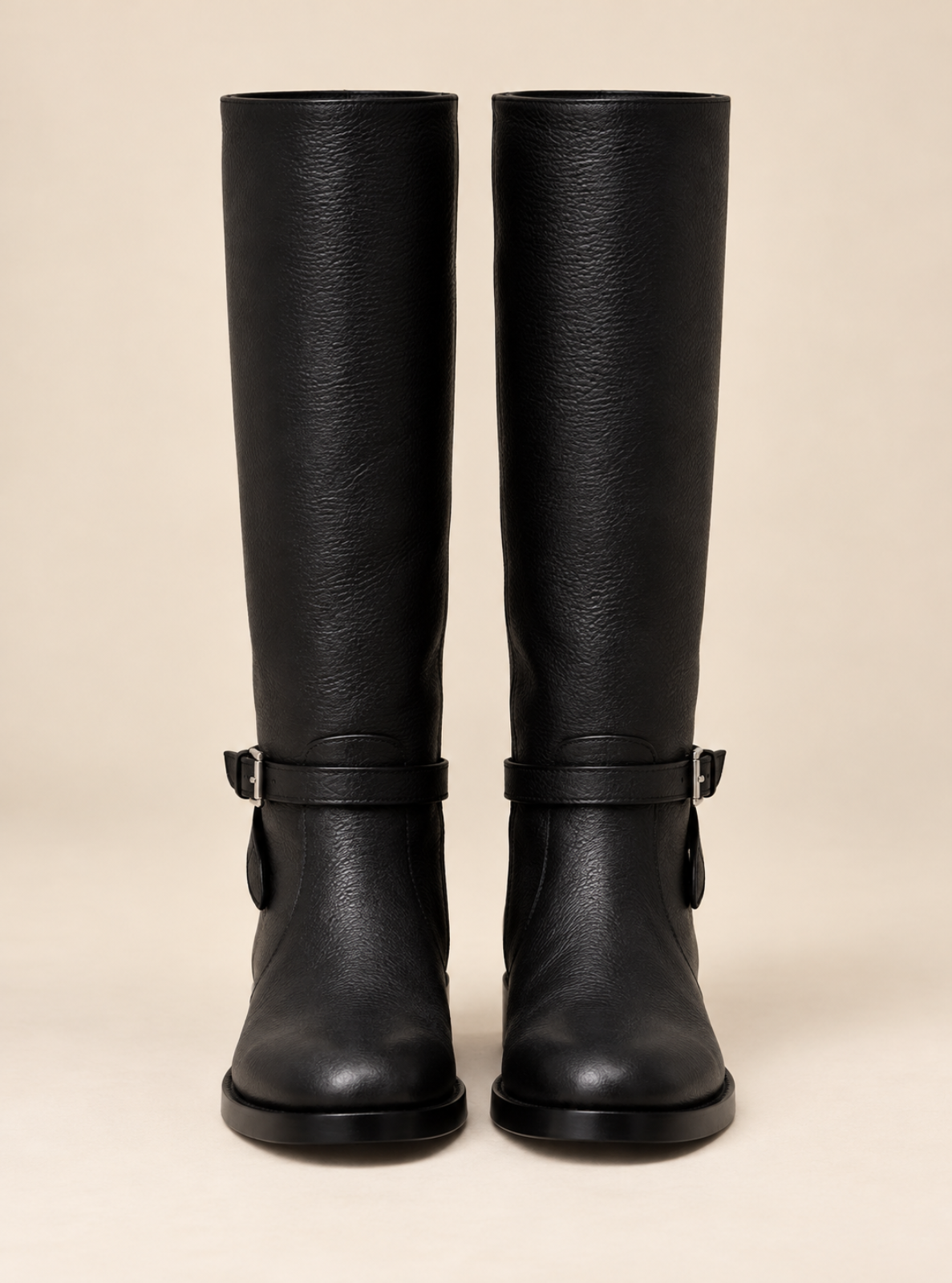 A pair of black leather knee-high riding boots with buckle straps around the lower shaft, shown against a solid beige background.