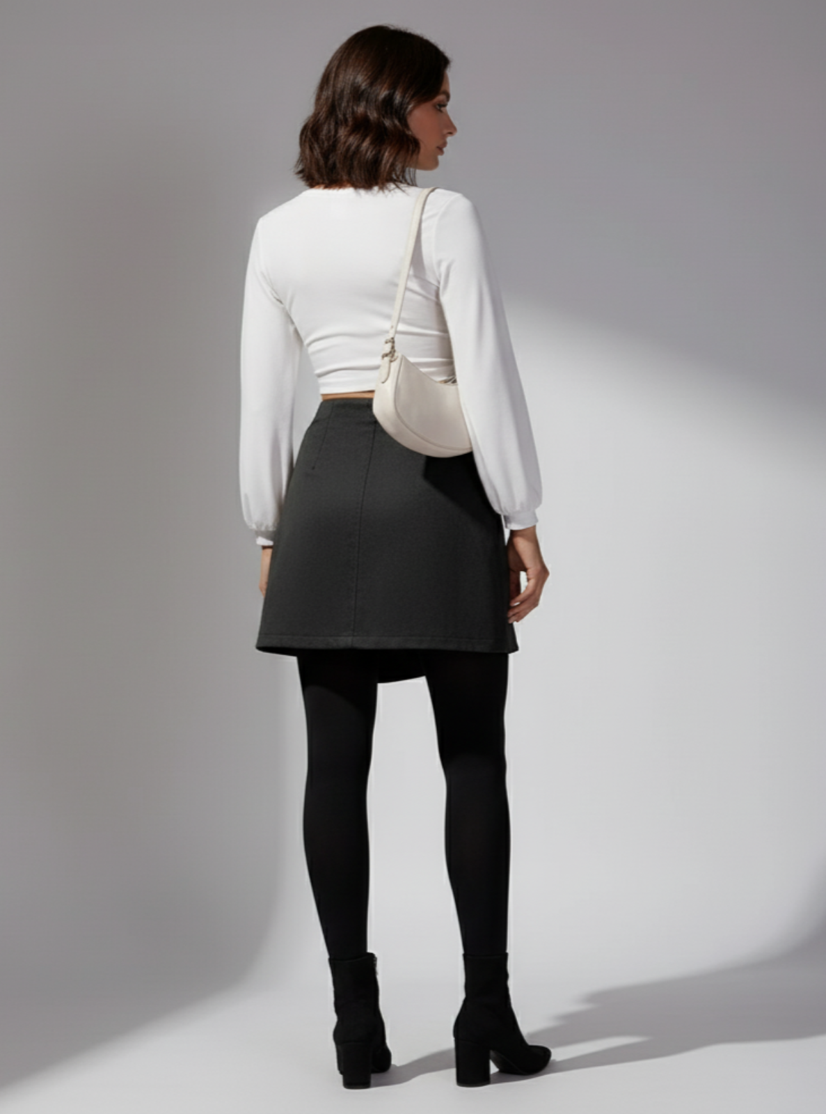 Woman wearing a white long-sleeve top, black skirt, and black tights against a gray background