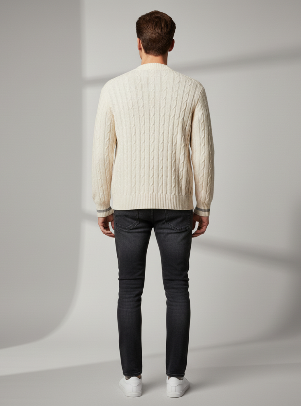 Man wearing a cream sweater and dark jeans standing against a plain background