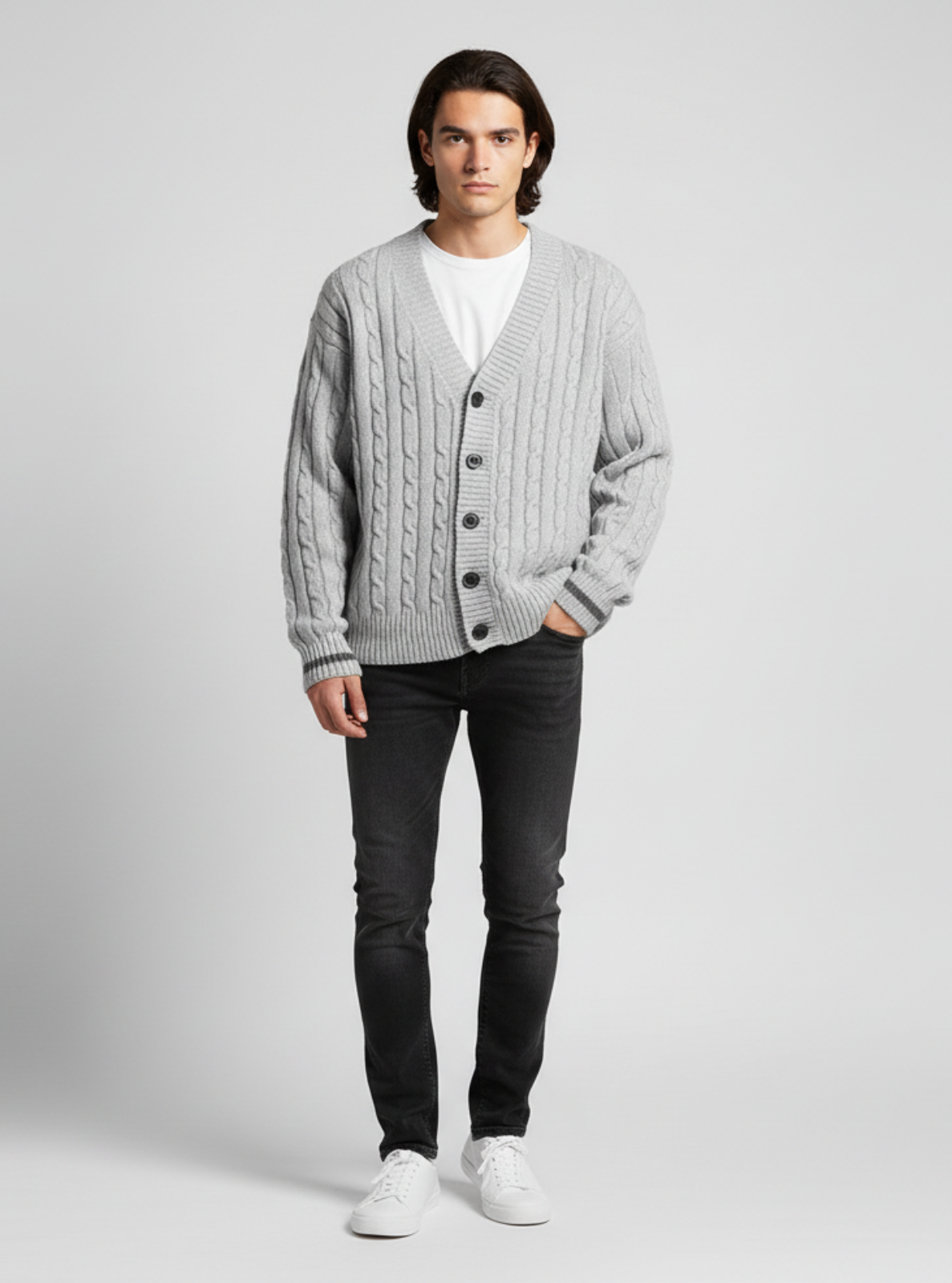 Man wearing a gray cable knit cardigan over a white shirt, paired with black pants and white sneakers on a light gray background.