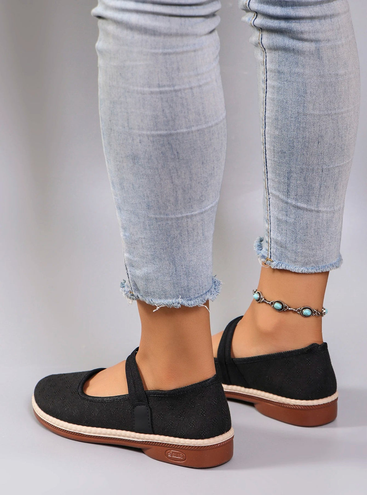 Black flat shoes with brown soles worn with light blue jeans on a neutral background