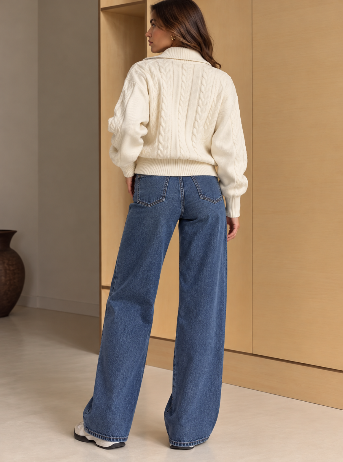 A woman is shown from behind wearing a cream cable-knit sweater and wide-leg blue jeans in a minimalist indoor setting with beige walls and wooden paneling.