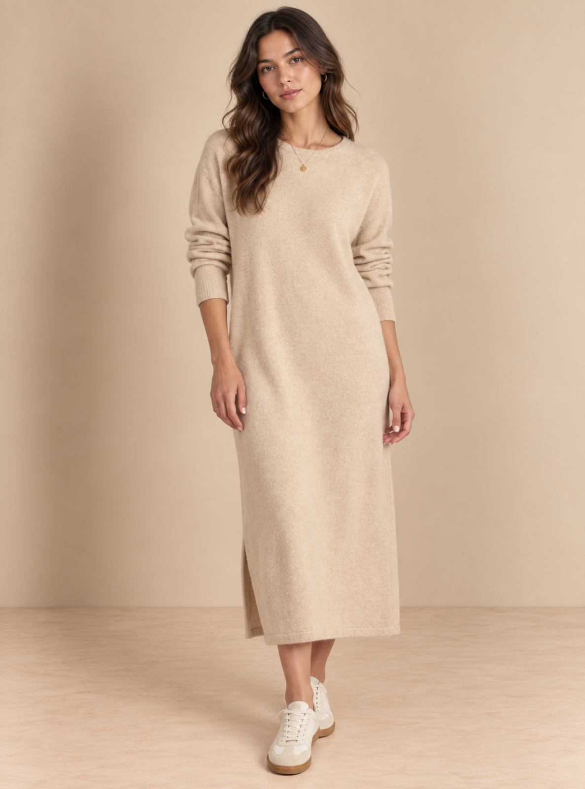 A woman modeling a long-sleeve beige knit midi dress with side slits, paired with white sneakers, against a warm beige studio backdrop.