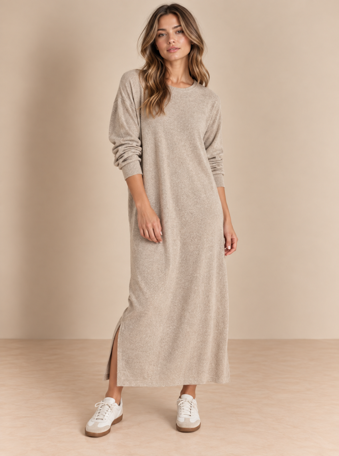 A woman modeling a long-sleeve, crew-neck maxi knit dress in a heathered light taupe-beige color. The dress features a side slit near the ankle and is styled with white sneakers against a neutral beige studio backdrop.