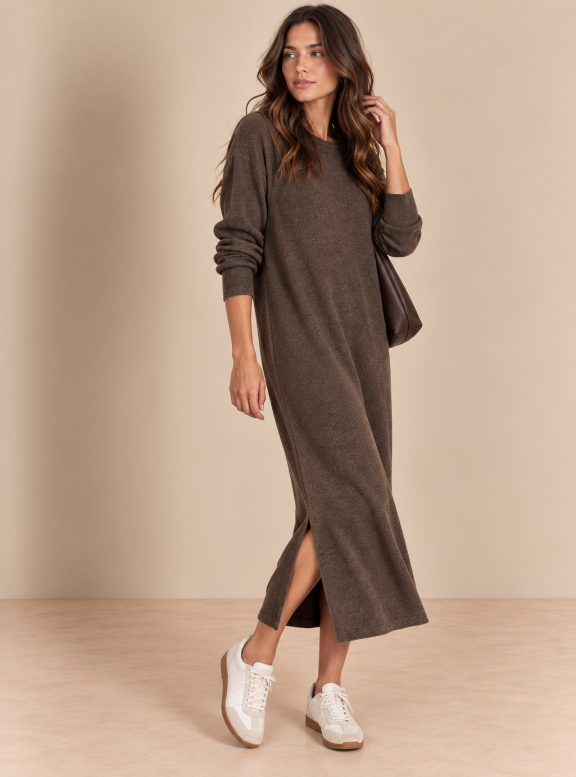 A woman models a long-sleeve, crewneck knit midi dress in a dark brown color, styled with white sneakers and a dark brown shoulder bag against a warm beige studio backdrop.