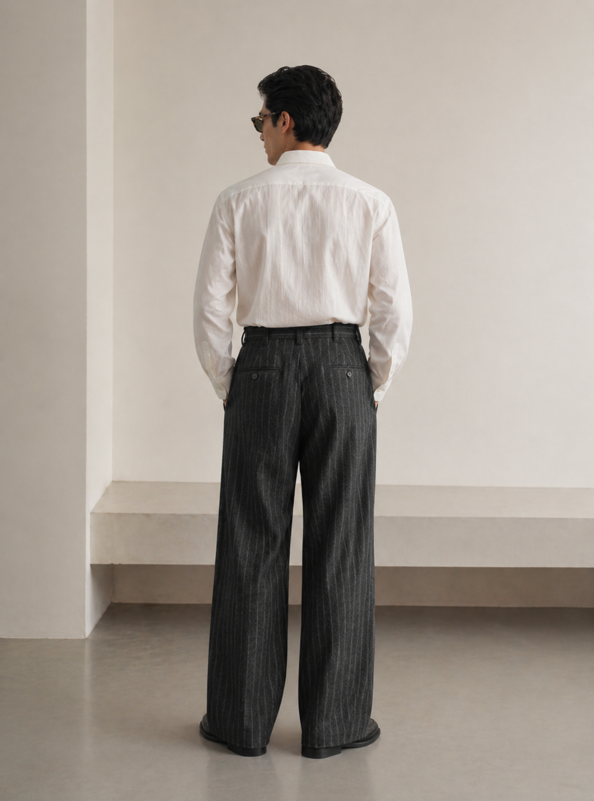 Man wearing a white shirt and dark pinstripe pants standing against a plain wall.