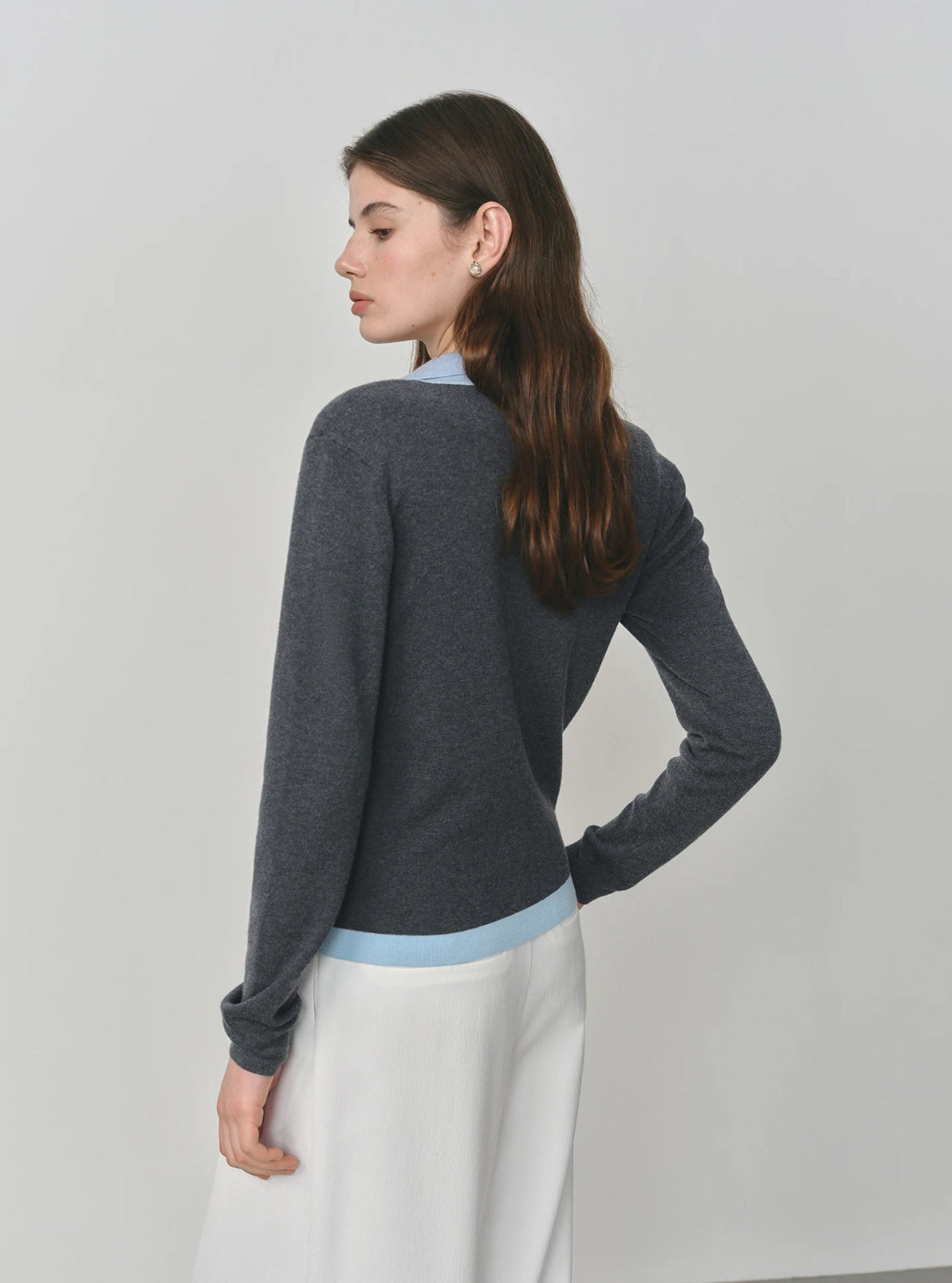 Woman wearing a gray sweater over a light blue shirt with white pants on a plain background