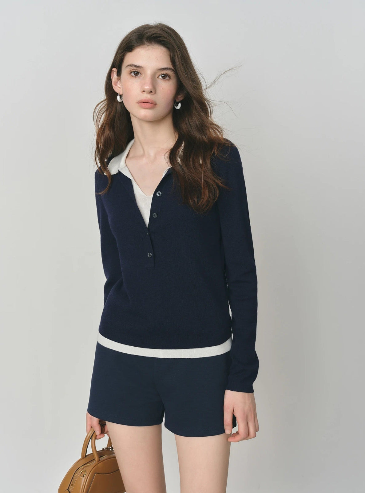 Woman wearing a navy blue cardigan over a white shirt with matching shorts, holding a brown bag against a plain background.