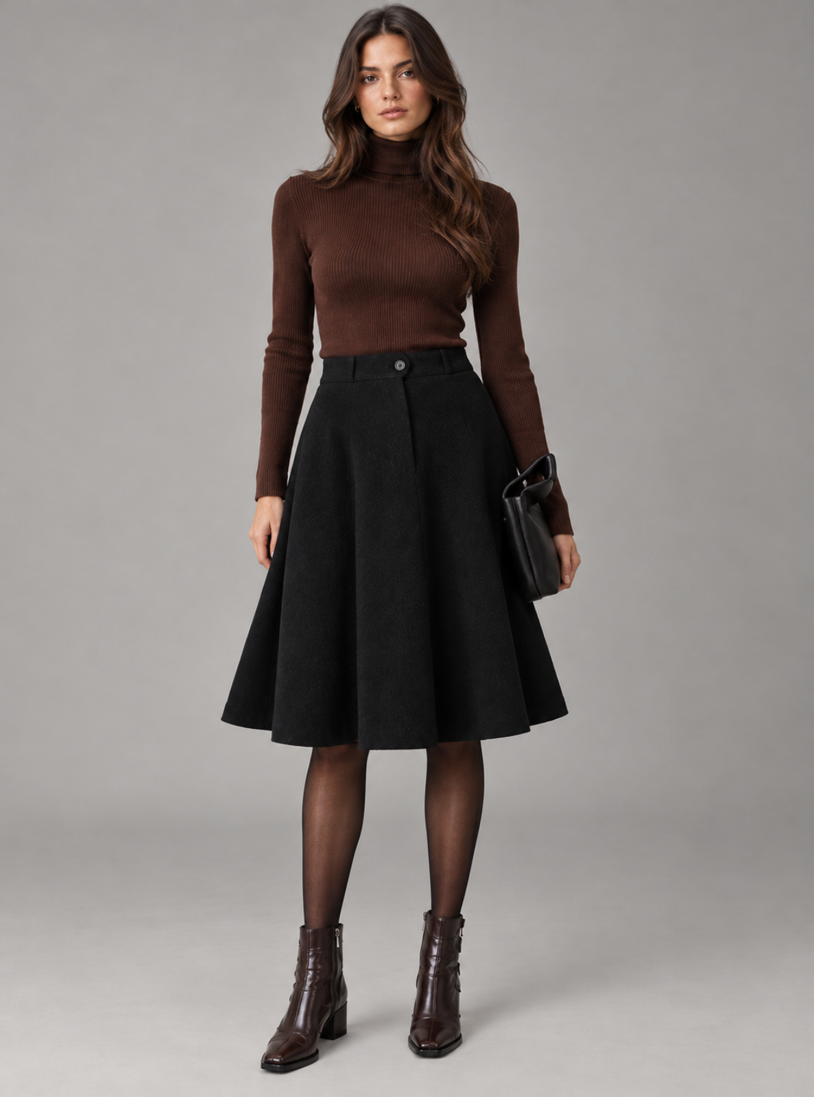 A woman modeling a brown ribbed turtleneck sweater, a black high-waisted A-line skirt, black tights, and dark brown ankle boots, holding a small black leather handbag against a solid gray studio background.