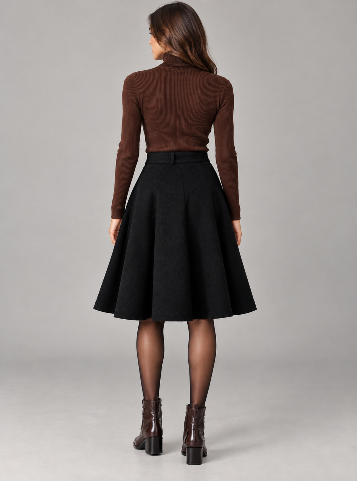 A woman shown from the back wearing a brown ribbed mock-neck long-sleeve top, a black high-waisted flared skirt, sheer black tights, and brown ankle boots, against a solid gray studio background.