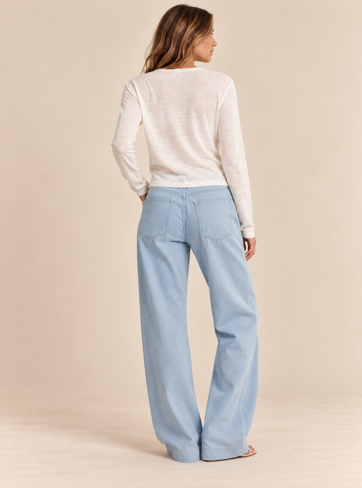 A woman shown from the back wearing a cream long-sleeve knit top and light blue wide-leg jeans, standing against a solid beige studio background.