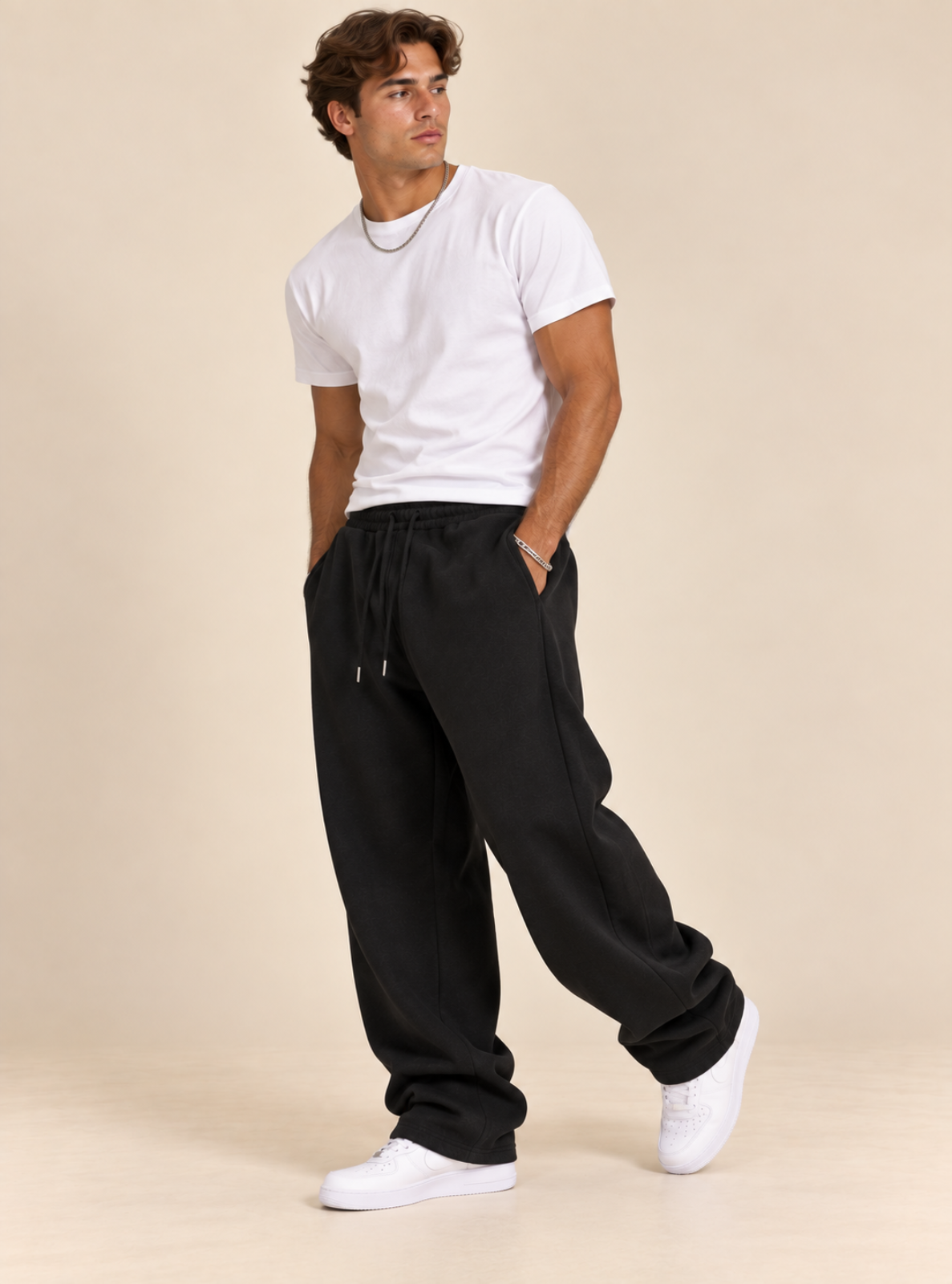 A man modeling black sweatpants with a white t-shirt and white sneakers against a solid beige background.