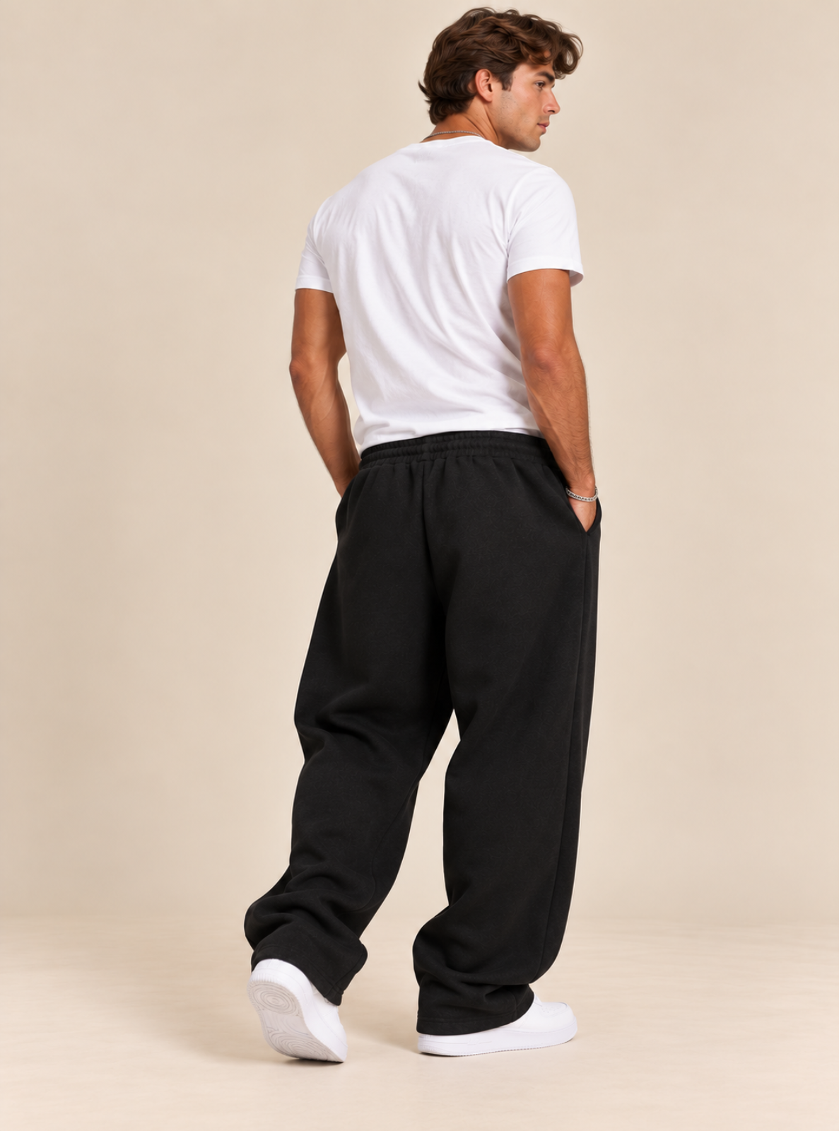 A man shown from the back wearing loose black sweatpants, a white t-shirt, and white sneakers, standing against a plain beige studio background.