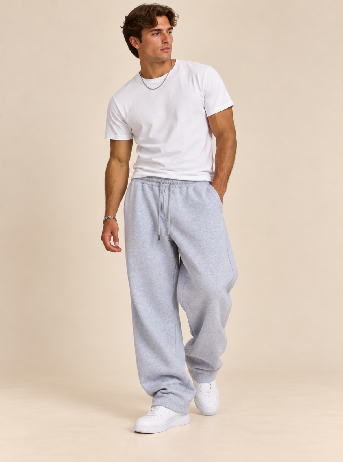 A man modeling a white crew-neck t-shirt and light gray heather sweatpants with white sneakers, standing against a solid beige studio background.