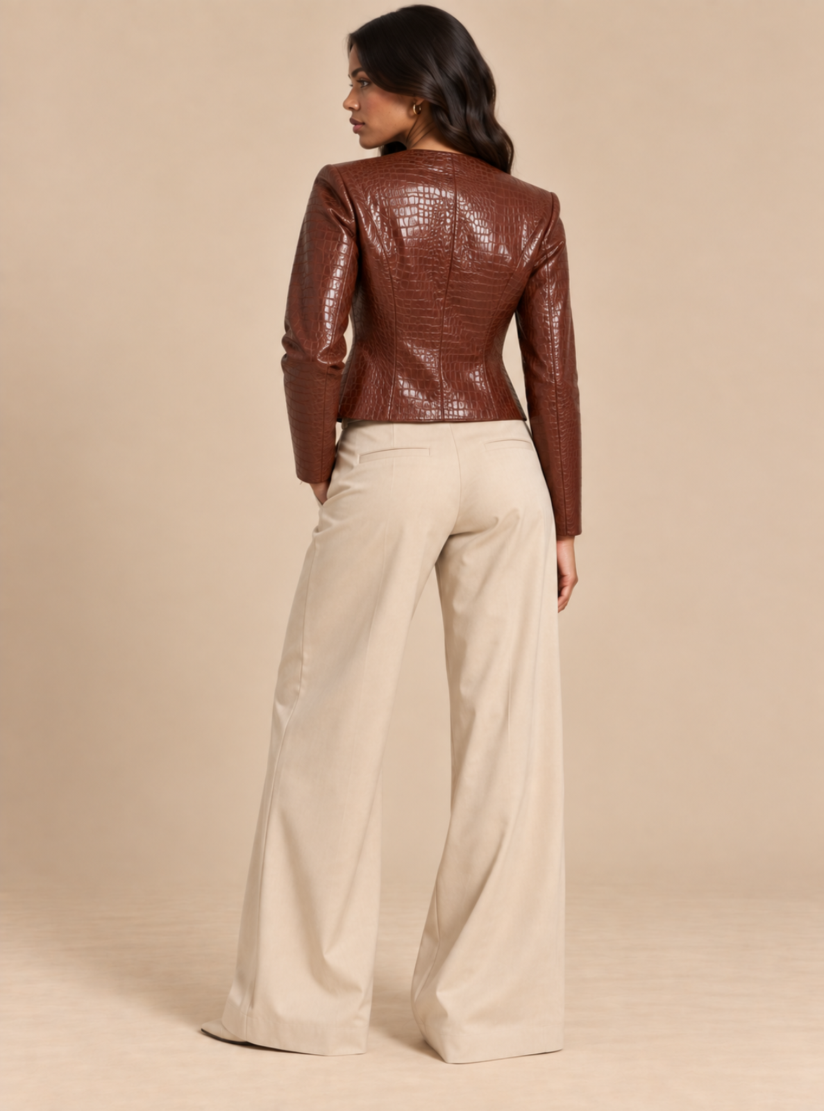 A woman shown from the back wearing a cropped brown faux-croc leather jacket and cream wide-leg trousers, standing against a solid beige studio background.