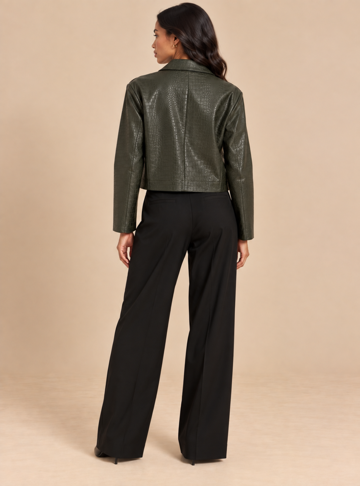 A woman shown from the back wearing a cropped, textured dark olive green faux-leather jacket and black wide-leg trousers, standing against a solid beige studio background.