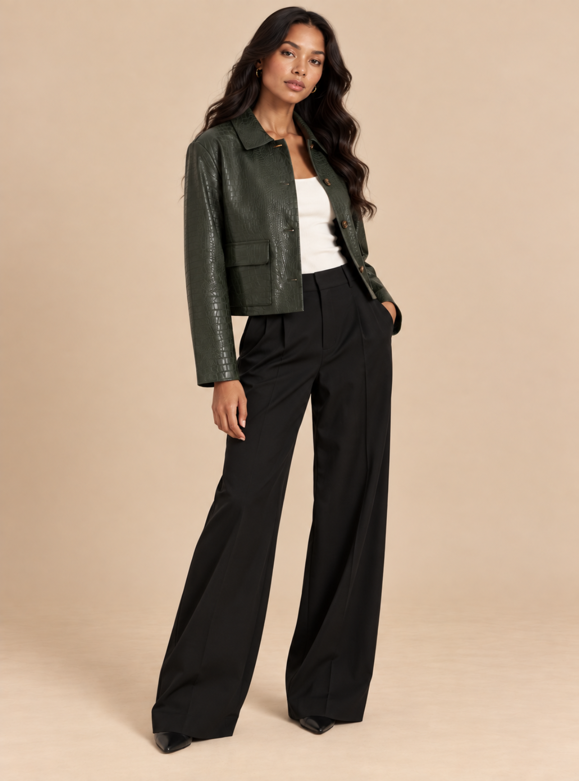 A woman models a dark green faux-leather jacket, styled with a white top and black wide-leg trousers, against a solid beige studio background.