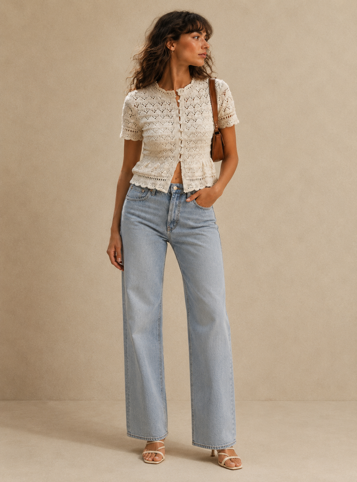 A woman stands against a plain beige studio backdrop wearing a cream lace short-sleeve cropped top, light blue wide-leg jeans, and beige heeled sandals, with a brown shoulder bag.