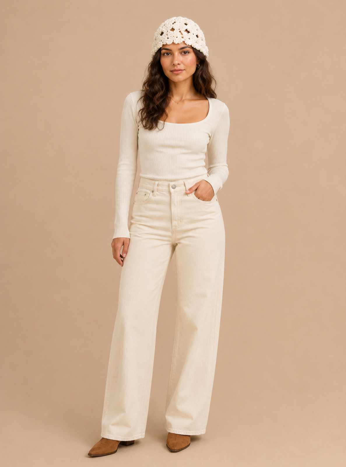 A woman stands against a plain beige studio background wearing a cream long-sleeve scoop-neck top, matching cream wide-leg jeans, a cream knit beanie, and brown pointed-toe flats.