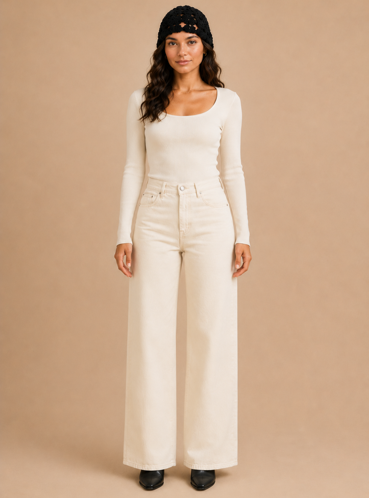 A woman stands against a plain beige studio background wearing a fitted long-sleeve scoop-neck top, high-waisted wide-leg pants, and a black beanie.