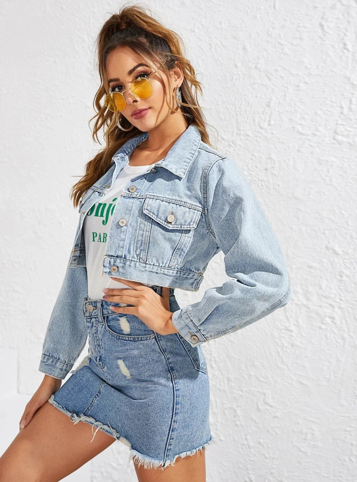 Woman wearing a denim jacket and shorts set with sunglasses against a white background