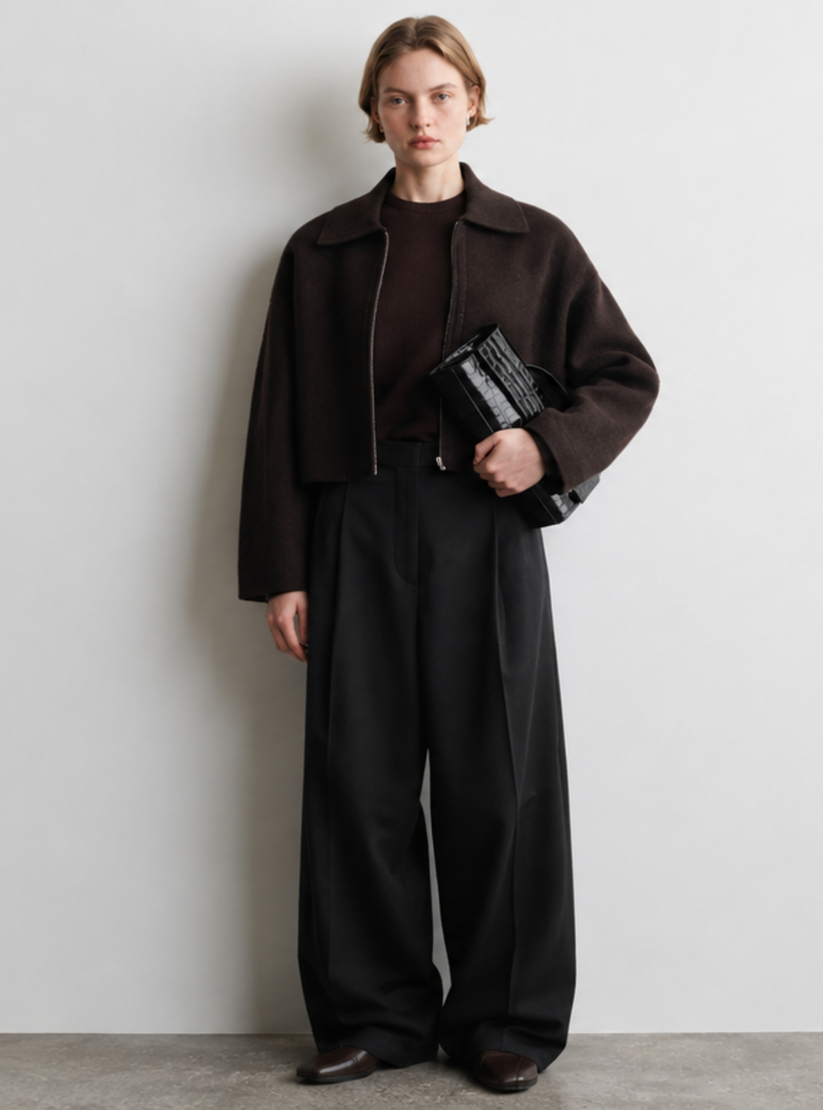 A model stands against a minimalist light gray wall wearing a dark brown cropped jacket over a dark top and wide-leg black trousers, holding a black clutch.