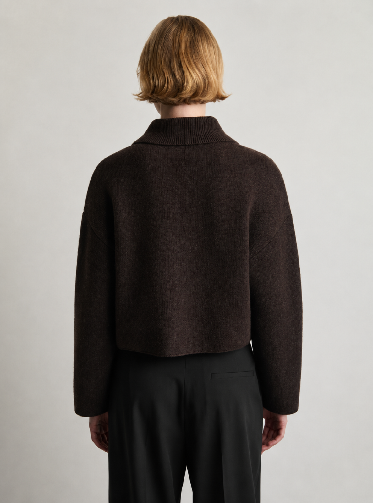 A model shown from the back wearing a dark brown cropped knit sweater with a mock neck and black trousers, set against a plain light gray studio background.