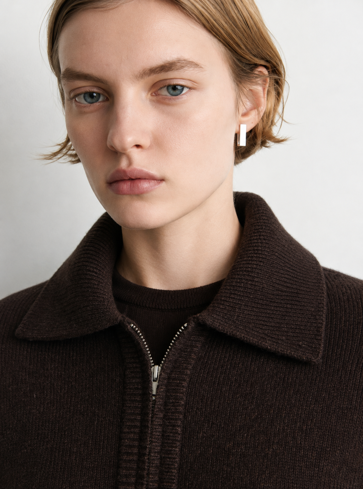Close-up of a model wearing a dark brown ribbed knit zip-up sweater with a wide collar over a matching dark brown top, set against a plain light gray background.