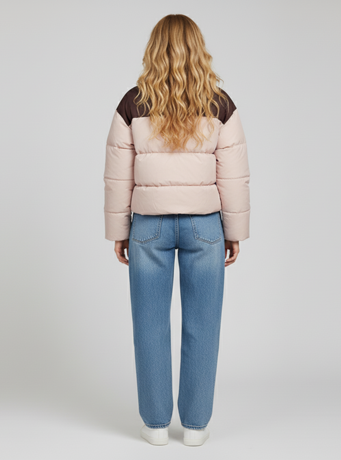 Person wearing a light pink puffer jacket with dark brown accents and blue jeans on a white background