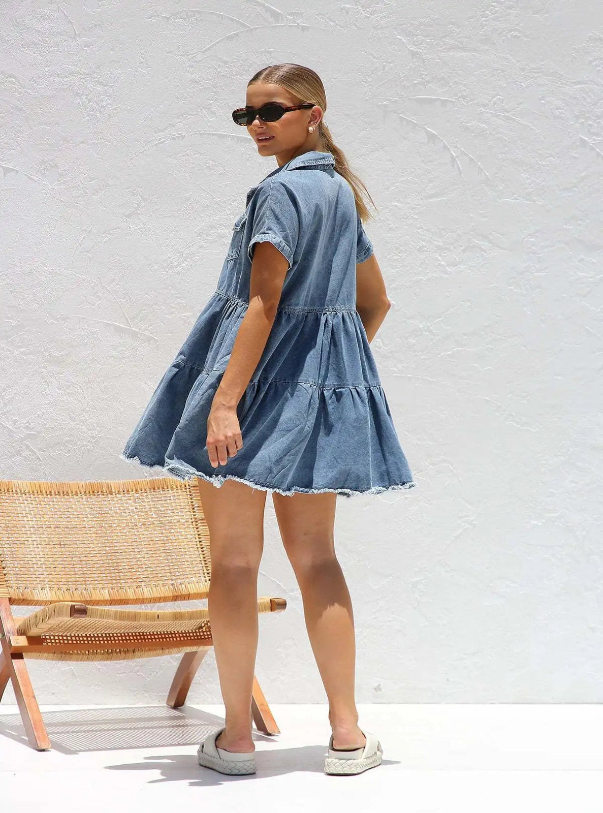 Woman wearing a denim dress standing against a white wall with a wicker chair.