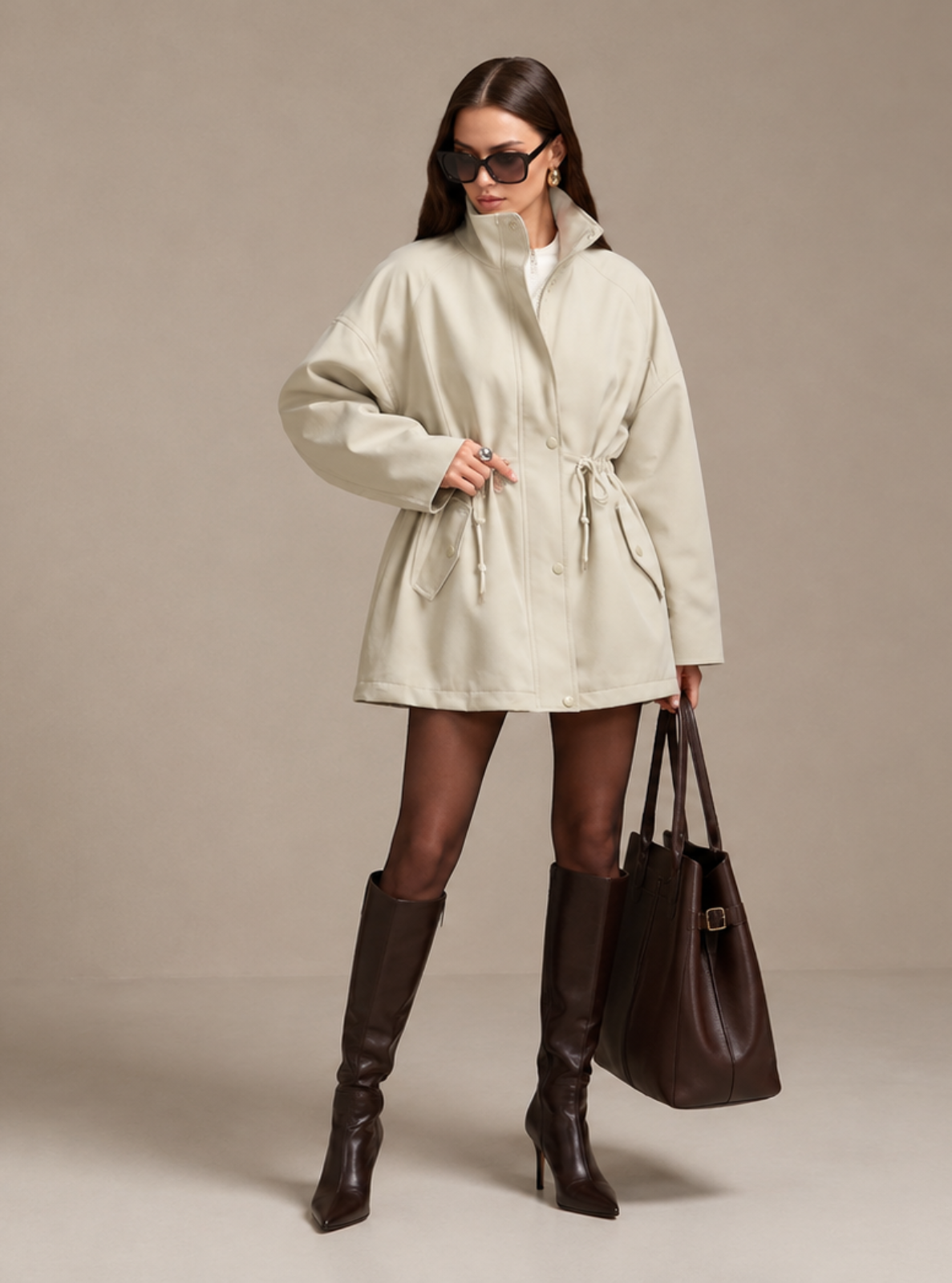 A woman modeling a cream-colored wrap coat with a tie waist, paired with dark brown knee-high boots and carrying a dark brown tote bag, set against a plain beige studio background.