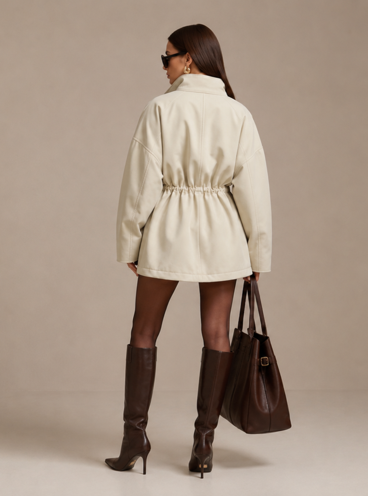 A model is shown from behind wearing a cream-colored belted coat, dark brown knee-high boots, and holding a dark brown leather tote bag against a solid beige studio background.