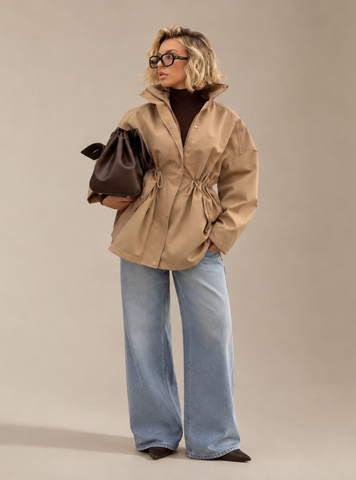 A woman stands against a plain beige studio background wearing a tan hooded jacket over a dark brown turtleneck, light blue wide-leg jeans, and dark shoes, while holding a dark brown handbag.