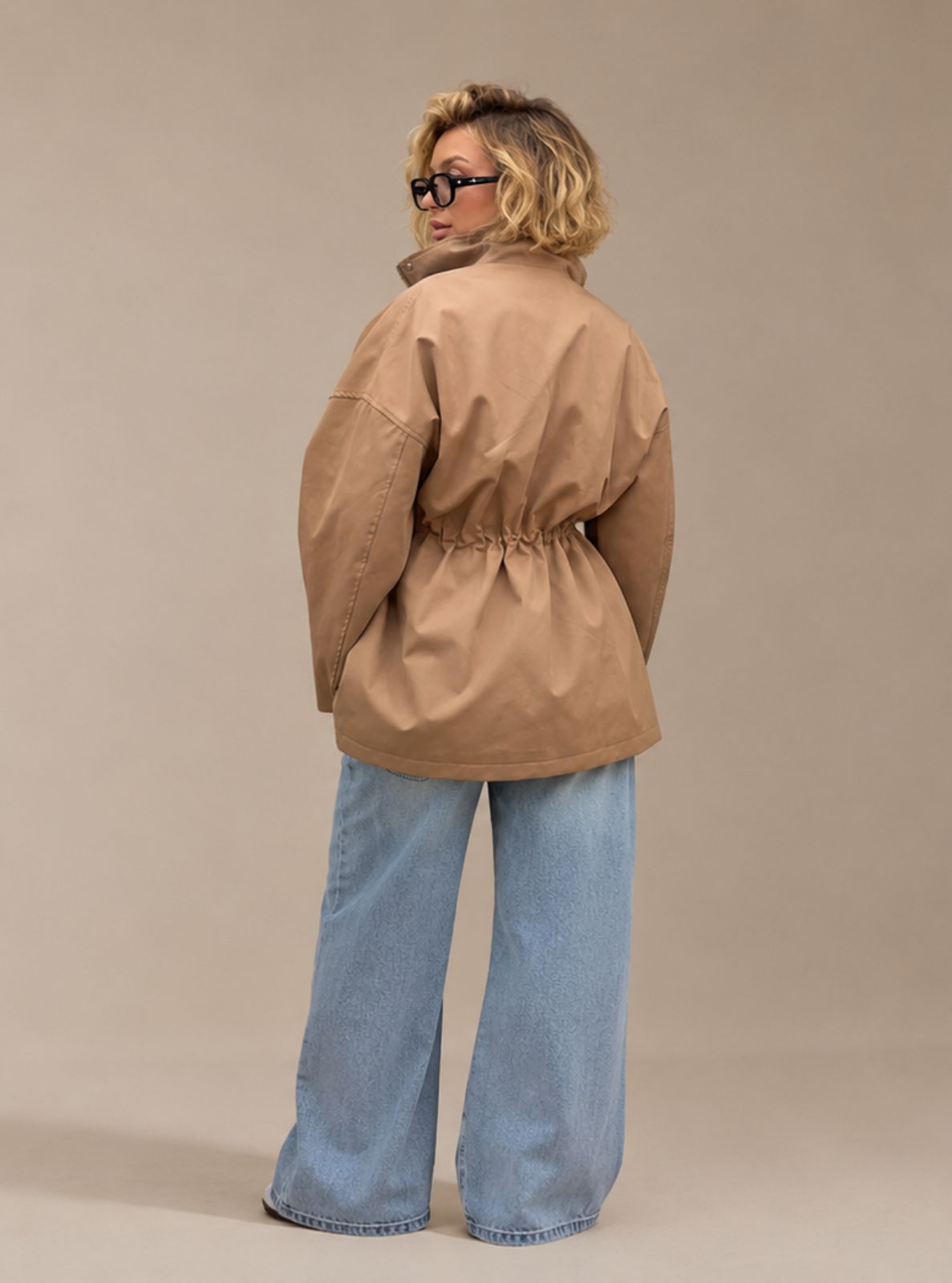 A woman with blonde hair and glasses, viewed from behind, wearing a tan hooded jacket with a drawstring waist and light blue wide-leg jeans, standing against a solid beige studio background.