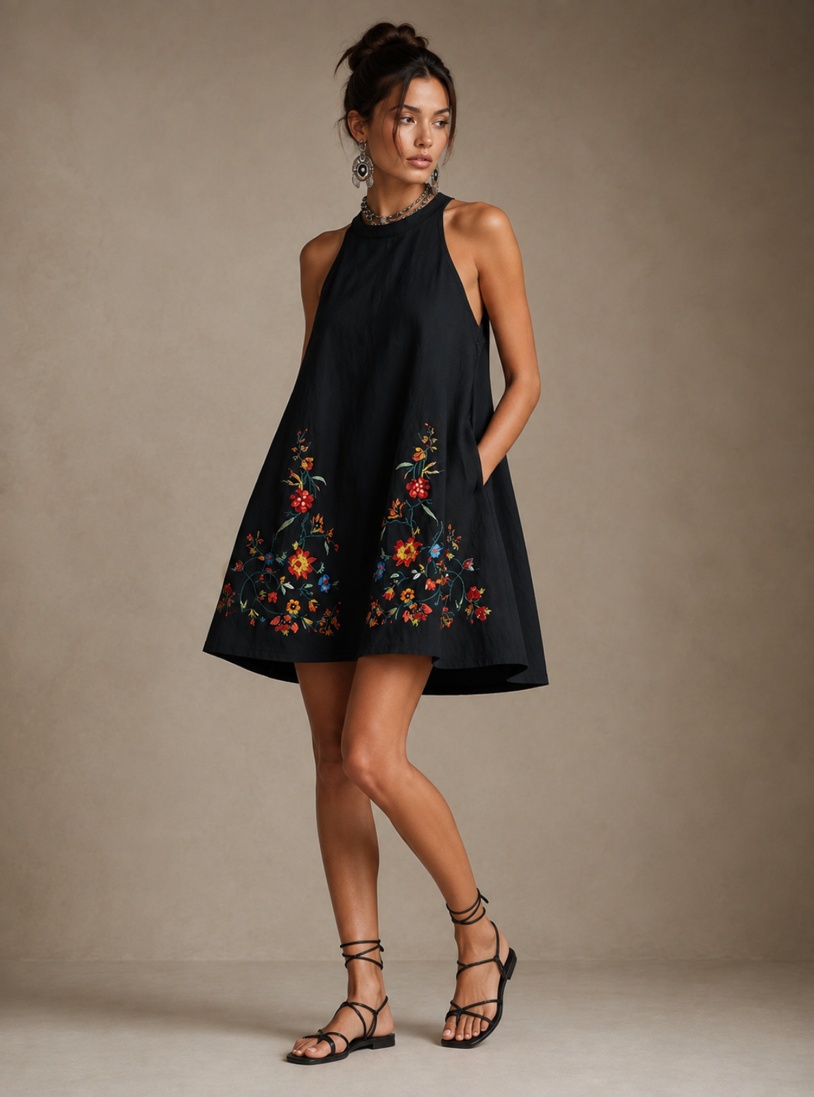 A woman models a sleeveless black halter mini dress with colorful floral embroidery near the hem, paired with black strappy flat sandals, against a neutral beige studio backdrop.