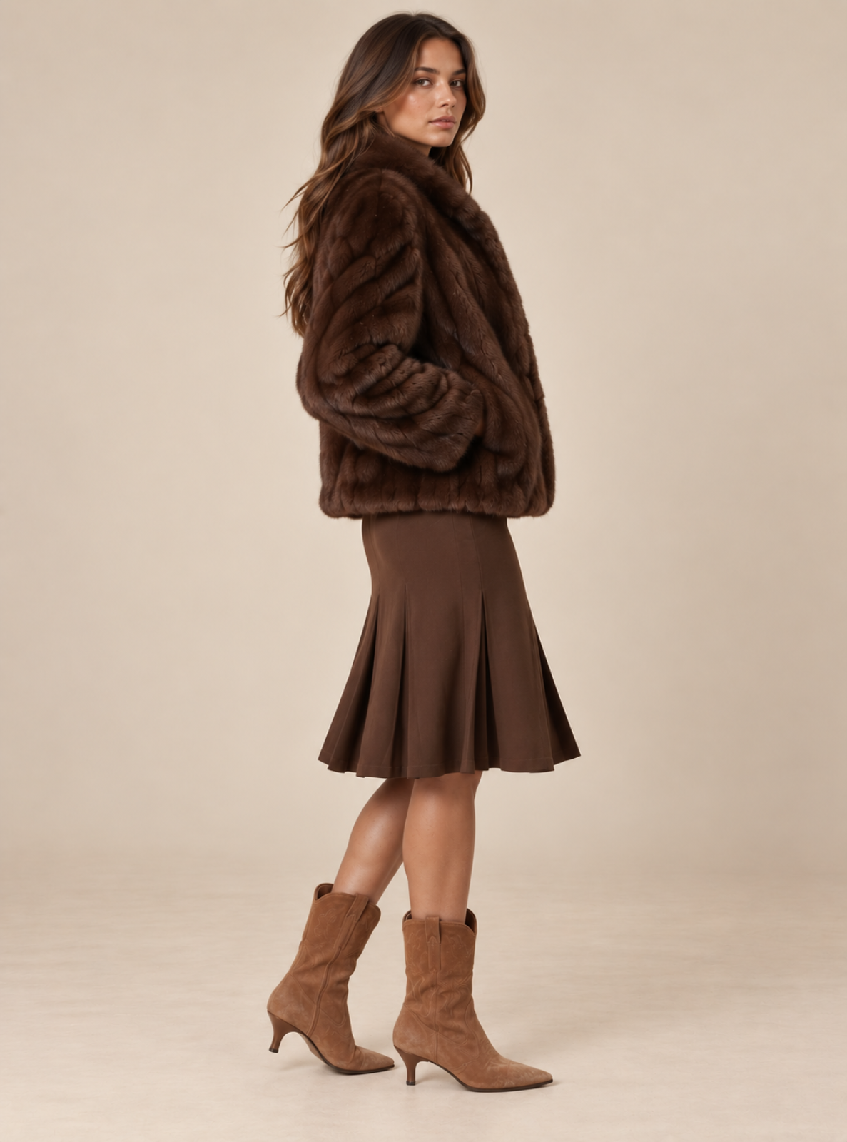 A woman modeling a dark brown faux fur jacket, a matching brown pleated midi skirt, and tan heeled ankle boots against a solid beige studio background.