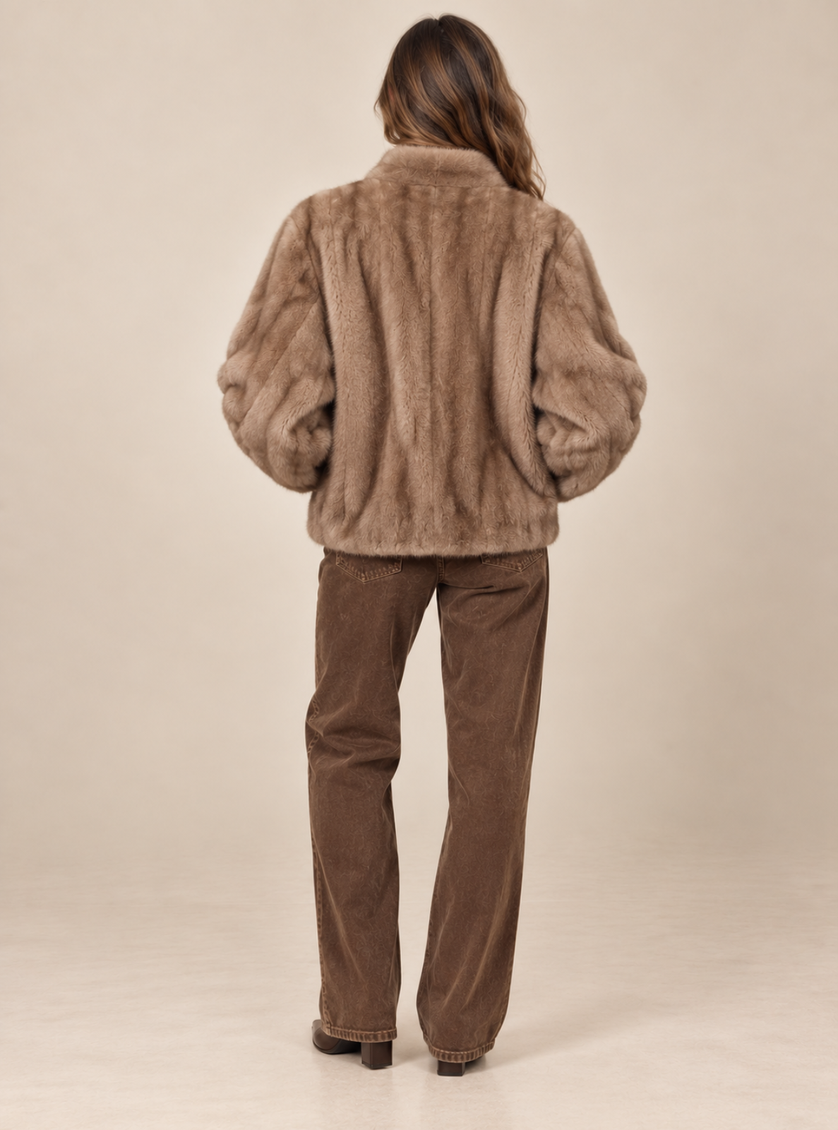 A woman viewed from behind wearing a brown faux fur jacket and brown corduroy pants against a plain beige studio background.