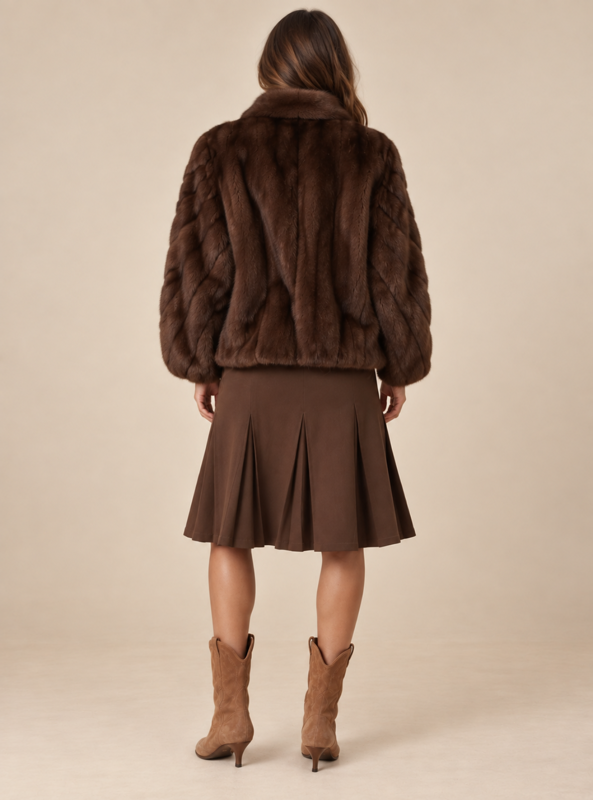 A woman shown from the back wearing a dark brown faux fur cropped jacket, a matching brown pleated skirt, and brown ankle boots, set against a plain beige studio background.