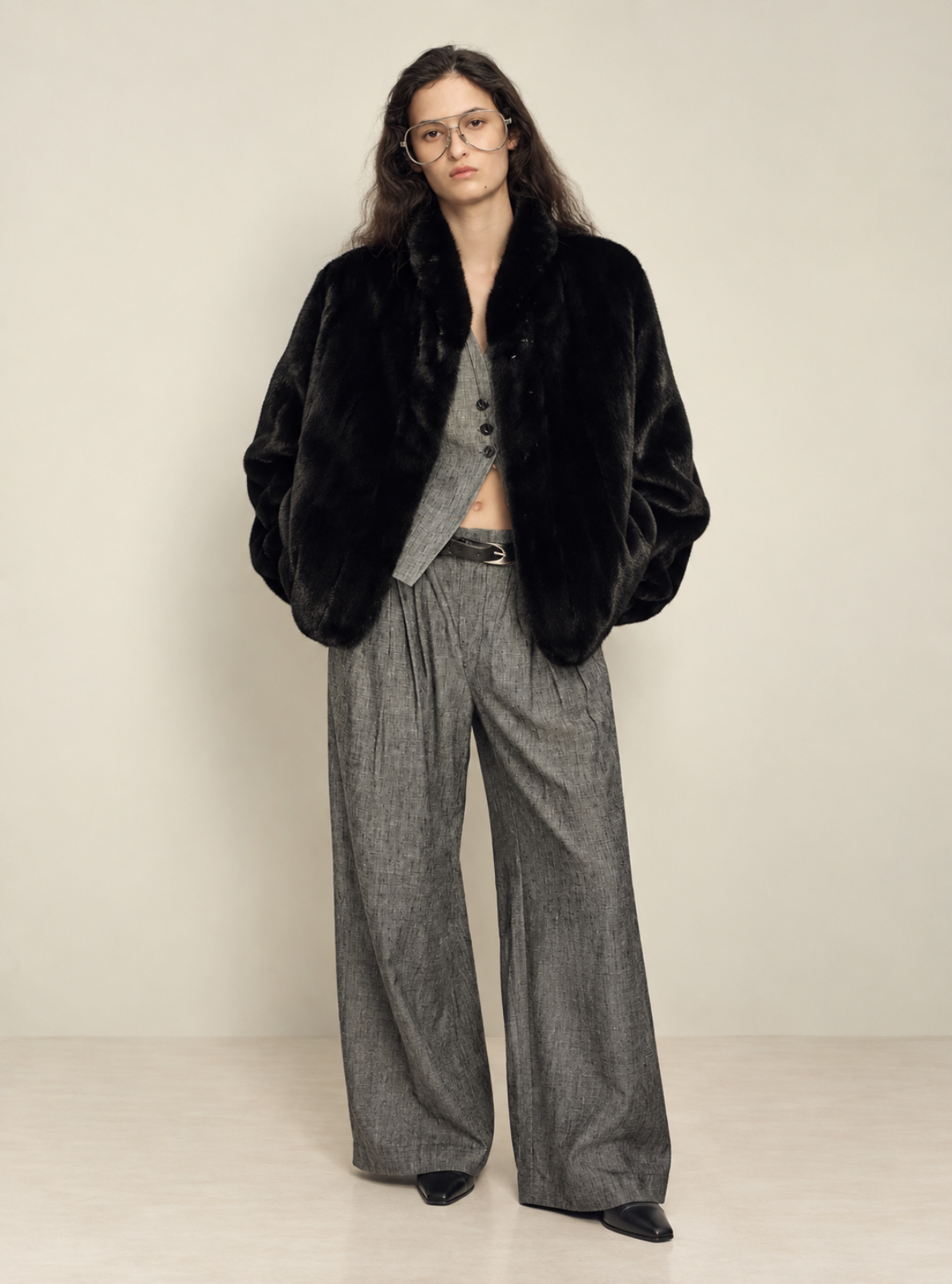 A model stands against a plain beige studio background wearing a black fur jacket over a gray cropped vest and matching gray wide-leg trousers.
